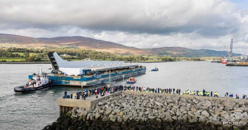Transport stalowej konstrukcji Narrow Water Bridge na barce w Warrenpoint, obserwowany przez mieszkańców zgromadzonych na falochronie.
